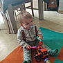 toddler, child, toy, pacifier, carpet, socks, suspenders, plaid_shirt, toy_tractor, rattle, floor, table, chair, indoor, home, playing, cute, baby, person, smiling
