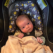 Alison is registered to the contest to win money with this photo: baby, infant, car_seat, blanket, floral_pattern, face, head, clothing, sitting, child, person, indoor, cute, newborn, soft_texture, hands, eyes, seatbelt, padded, portrait