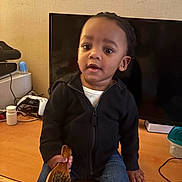 Angel is registered to the contest to win money with this photo: child, toddler, hairbrush, desk, television, black_jacket, jeans, indoor, curious, person, furniture, technology, container, wood, home, casual_clothing, sitting, young_child, curly_hair, expression