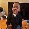 child, toddler, hairbrush, desk, television, black_jacket, jeans, indoor, curious, person, furniture, technology, container, wood, home, casual_clothing, sitting, young_child, curly_hair, expression