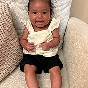 Ariella joined the competition — help win amazing prizes! baby, child, smiling, sitting, bow, hair_accessory, black_shorts, white_top, sandals, footwear, chair, cushion, indoor, cute, happy, portrait, person, infant, skin, furniture