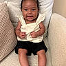 baby, child, smiling, sitting, bow, hair_accessory, black_shorts, white_top, sandals, footwear, chair, cushion, indoor, cute, happy, portrait, person, infant, skin, furniture