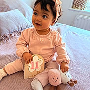 Lyna participe au concours pour gagner de l'argent avec cette photo : baby, girl, infant, pink_clothing, headband, milestone_sign, plush_toys, bed, blanket, pillow, natural_light, indoors, smiling_face, sitting, cute, child, soft_texture, window, decor, happy