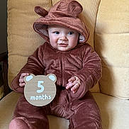 Nathan is registered to the contest to win money with this photo: baby, child, infant, onesie, hood, ears, bear_costume, sign, wooden_sign, months, cushion, chair, furniture, cute, smiling, sitting, indoors, portrait, soft_texture, clothing