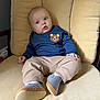 baby, child, infant, sweater, bear_patch, pants, socks, shoe, chair, cushion, indoor, furniture, person, young_child, cute, relaxed, seated, looking_away, cozy, home