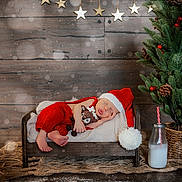 Kade joined the competition — help win amazing prizes! newborn, baby, sleeping, santa_hat, red_clothing, plush_toy, wooden_bed, blanket, christmas_tree, pine_cones, red_berries, milk_bottle, striped_straw, wooden_background, golden_stars, rug, holiday_theme, festive, indoors, cozy