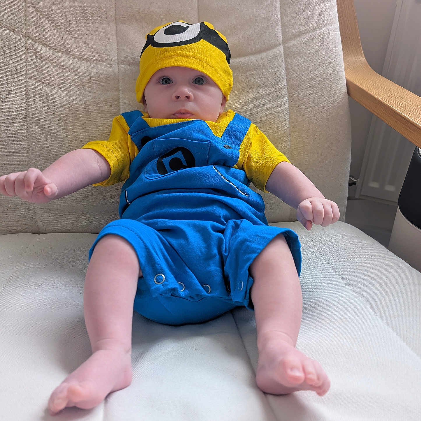 Reuben joined the competition — help win amazing prizes! arms, baby, blue_overalls, child, clothing, cushioned_chair, cute, expression, face, furniture, home, indoor, infant, legs, minion_hat, person, portrait, relaxing, seated, yellow_hat