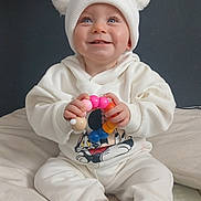 Eliza is registered to the contest to win money with this photo: baby, child, white_clothing, hat, pom_pom, toy, colorful, smiling, blue_eyes, sitting, indoors, pillow, cute, happy, face, hands, sock, floor_mat, cozy, portrait