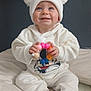 baby, child, white_clothing, hat, pom_pom, toy, colorful, smiling, blue_eyes, sitting, indoors, pillow, cute, happy, face, hands, sock, floor_mat, cozy, portrait