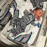 Damon is registered to the contest to win money with this photo: newborn, baby, hospital_bassinet, blanket, clothing, pacifier, infant, person, cute, lying_down, footwear, bed, striped_blanket, socks, hospital, cute_outfit, mustache_print, alert, small_hands, indoors