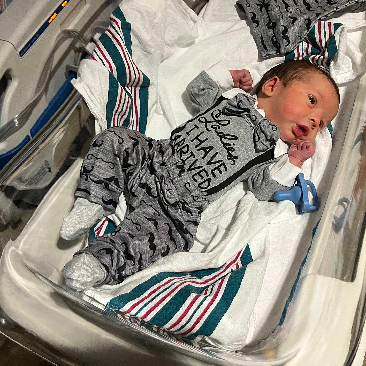 Damon is registered to the contest to win money with this photo: alert, baby, bed, blanket, clothing, cute, cute_outfit, footwear, hospital, hospital_bassinet, indoors, infant, lying_down, mustache_print, newborn, pacifier, person, small_hands, socks, striped_blanket