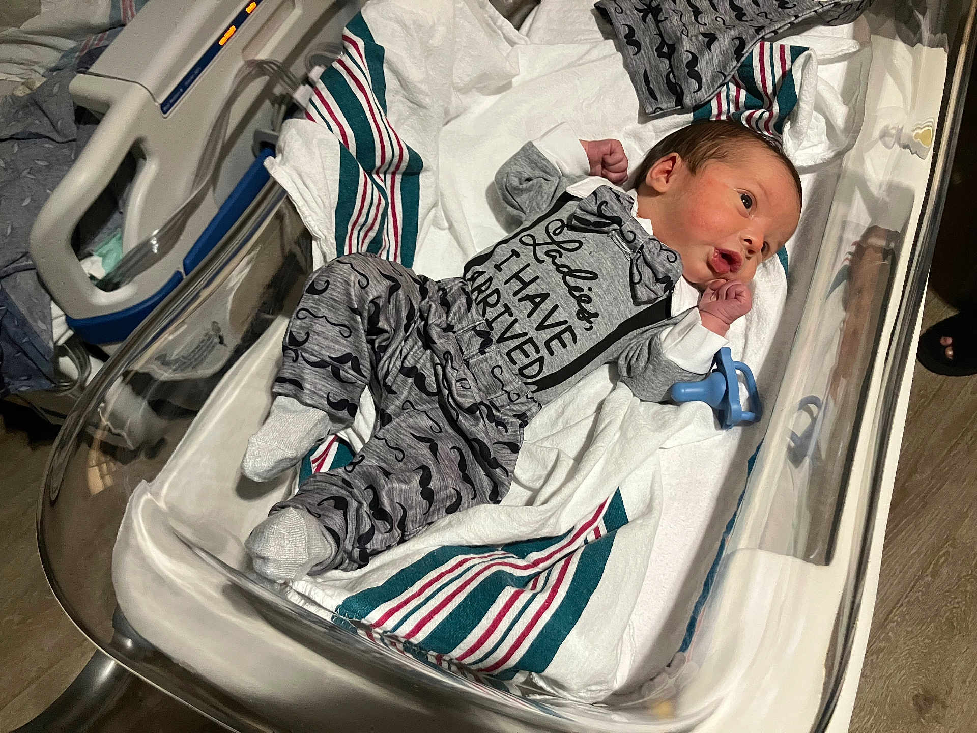 Damon is registered to the contest to win money with this photo: newborn, baby, hospital_bassinet, blanket, clothing, pacifier, infant, person, cute, lying_down, footwear, bed, striped_blanket, socks, hospital, cute_outfit, mustache_print, alert, small_hands, indoors