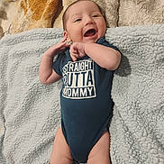 Jose is registered to the contest to win money with this photo: baby, infant, smiling, onesie, text_clothing, blanket, soft_texture, lying_down, happy, cute, child, person, indoor, newborn, skin, hand, face, portrait, clothing, playful