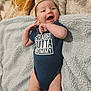 baby, infant, smiling, onesie, text_clothing, blanket, soft_texture, lying_down, happy, cute, child, person, indoor, newborn, skin, hand, face, portrait, clothing, playful
