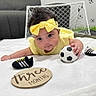 baby, infant, yellow_clothing, headband, soccer_ball, toy_shoes, mini_goal, white_blanket, plaque, three_months, indoor, floor, child, cute, small, playing, sports_theme, toddler, portrait, expression