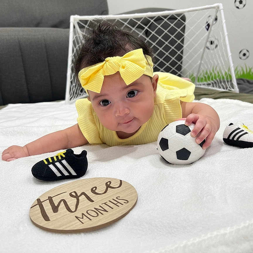 Sofia joined the competition — help win amazing prizes! baby, infant, yellow_clothing, headband, soccer_ball, toy_shoes, mini_goal, white_blanket, plaque, three_months, indoor, floor, child, cute, small, playing, sports_theme, toddler, portrait, expression