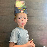 Joseph is registered to the contest to win money with this photo: child, boy, box, headwear, snack, gray_shirt, wooden_wall, table, number_38, indoor, playful, face, light_skin, short_hair, casual_clothing, holding, standing, portrait, expression, cheeky