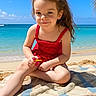 child, girl, beach, ocean, sand, towel, red_dress, polka_dot, smile, sunny, outdoor, summer, water, sky, waves, happy, sitting, playful, vacation, nature
