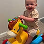 baby, child, toy, horse, indoor, carpet, smiling, happy, toddler, play, colorful, plastic, wheels, clothing, shorts, shirt, hand, foot, person, window