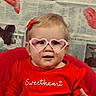 toddler, child, glasses, heart_shaped_glasses, red_sweater, sweetheart_text, newspaper_background, decorative_hearts, red_hair_clip, indoor, portrait, cute, smiling, baby, seat, pink, red, face, hand, fashion