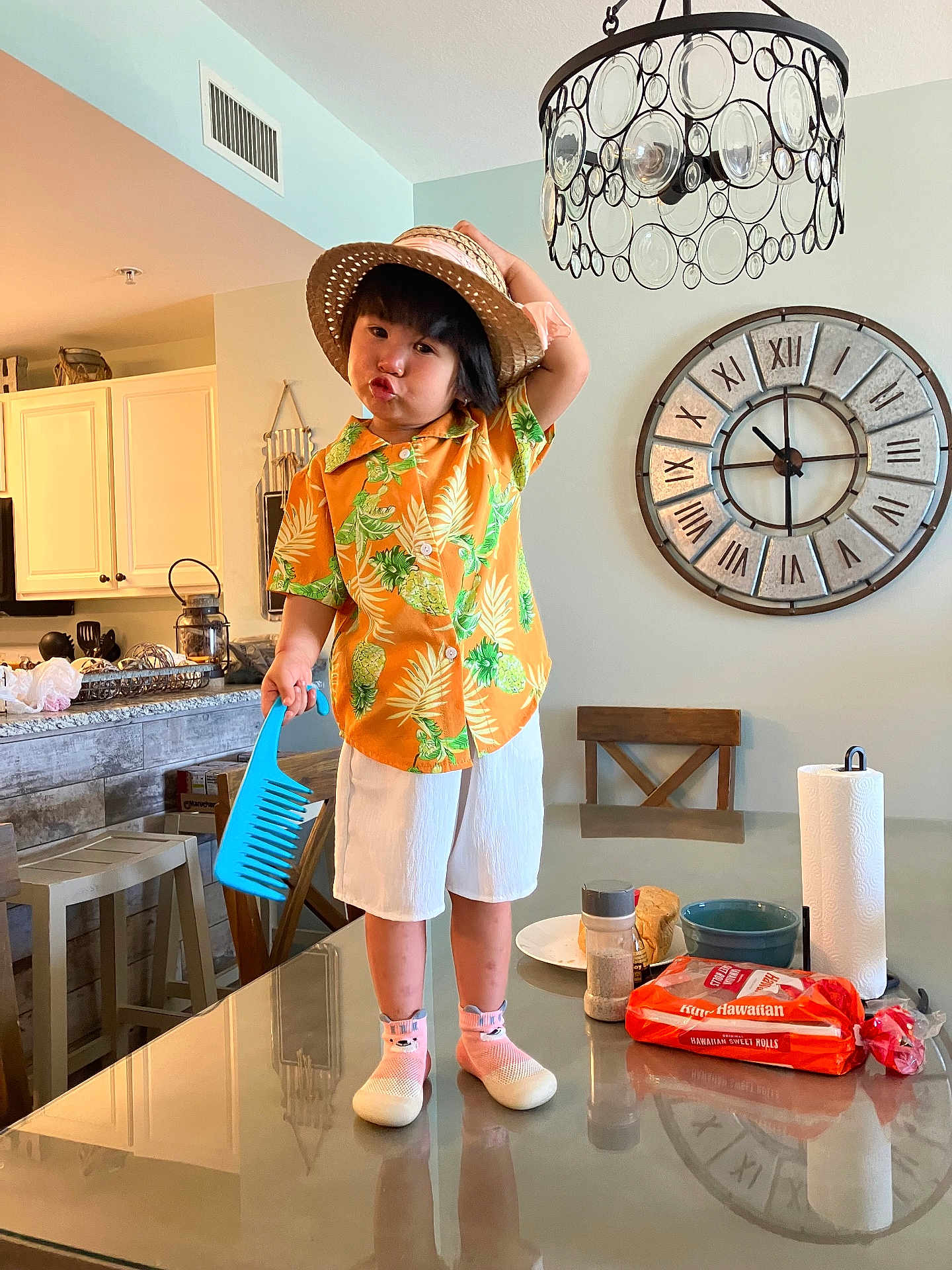 Tesla Lam Tran is registered to the contest to win money with this photo: child, person, table, hat, comb, orange_shirt, white_shorts, pink_socks, shoes, kitchen, clock, chair, glass_table, chandelier, food, bread, paper_towel, cup, counter, playful