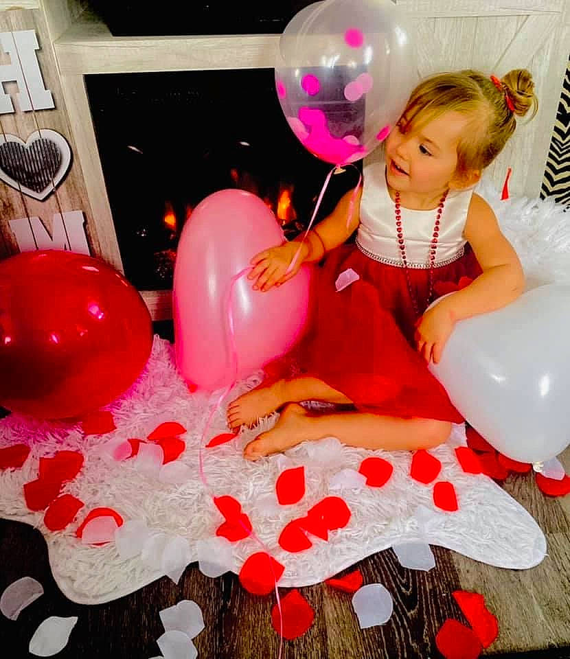 Mia is registered to the contest to win money with this photo: balloon, dress, event, facial_expression, fun, happy, leg, leisure, light, magenta, material_property, organ, party_supply, person, pink, red, sitting, thigh, toddler, toy