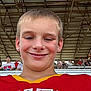 child, boy, smile, earrings, short_hair, red_jersey, sportswear, stadium, spectators, metal_roof, crowd, outdoor, daylight, casual, portrait, young, happy, event, people, seated