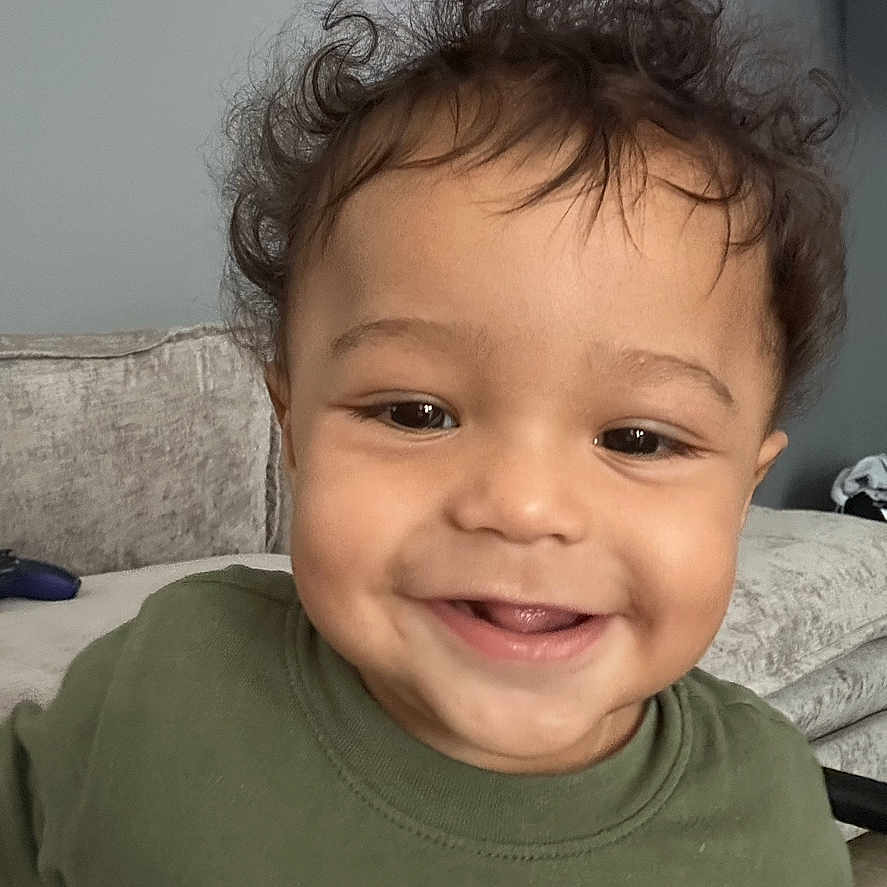 Kioni is registered to the contest to win money with this photo: baby, child, closeup, couch, curly_hair, cute, expression, face, green_shirt, happy, head, indoor, living_room, person, portrait, seated, skin, smiling, toddler, young_child