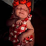 baby, sleeping, red_headband, rose_headband, checkered_dress, infant, child, person, hand, arm, dark_background, indoor, cute, peaceful, newborn, small_hands, soft_light, portrait, wearing, adorable
