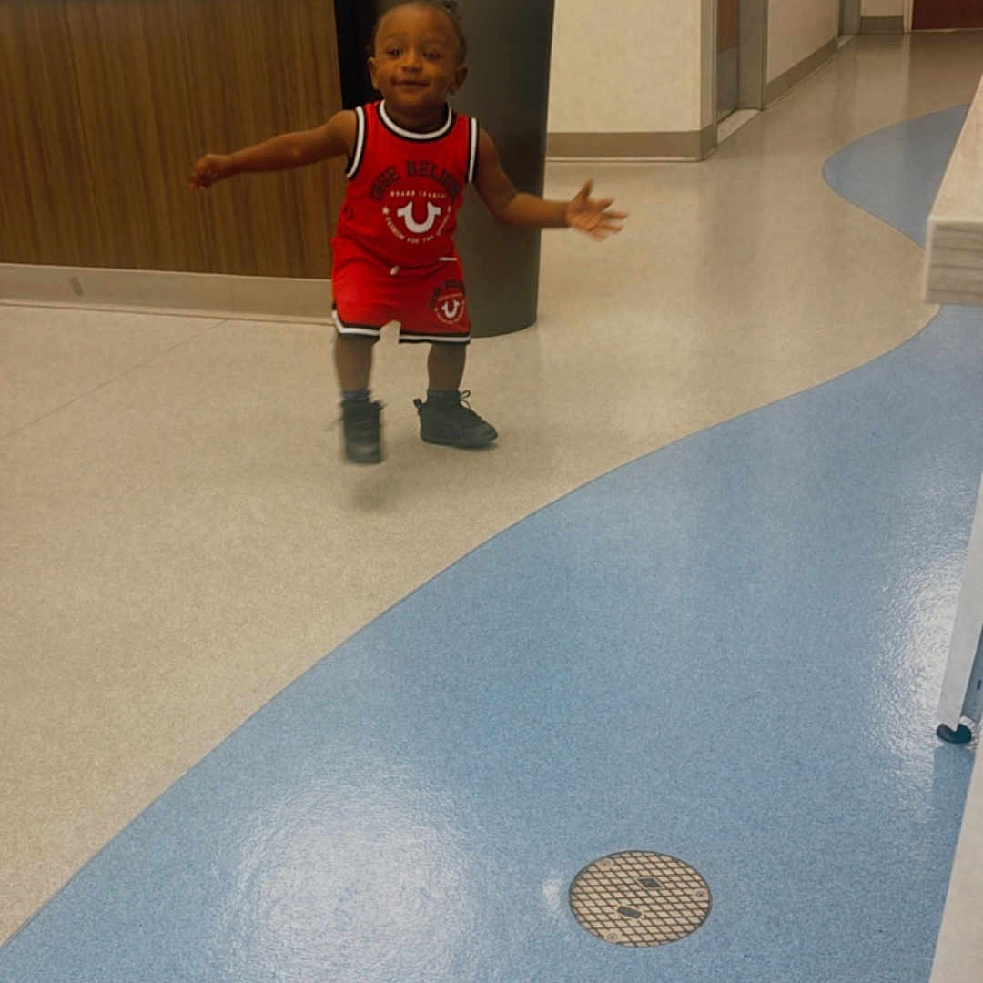 Deshawn joined the competition — help win amazing prizes! child, toddler, red_clothing, hallway, indoor, floor, plant, pot, door, smiling, person, shoes, wall, decor, motion, young_child, curved_floor_pattern, light, small_person, happy
