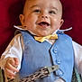 accessory, baby, blue_vest, child, closeup, clothing, cute, face, hand, happy, indoor, infant, lying_down, patterned_strap, person, portrait, red_background, smiling, white_shirt, yellow_bow_tie