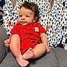 baby, baby_clothing, bare_feet, blanket, child, couch, curly_hair, face, gray_fabric, happy, home, indoor, infant, patterned_blanket, photography, portrait, red_romper, sitting, smiling, toes