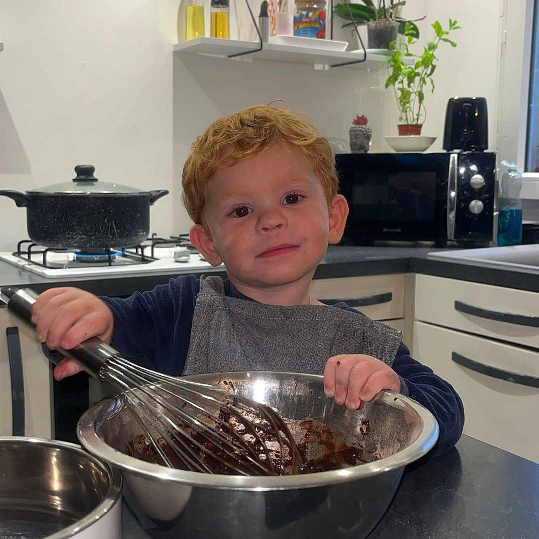 Mayrone participe au concours pour gagner de l'argent avec cette photo : apron, child, chocolate, cooking, countertop, cupboard, face, food_preparation, indoor, kitchen, microwave, mixing_bowl, oven, person, plants, pots, red_hair, smile, toddler, whisk