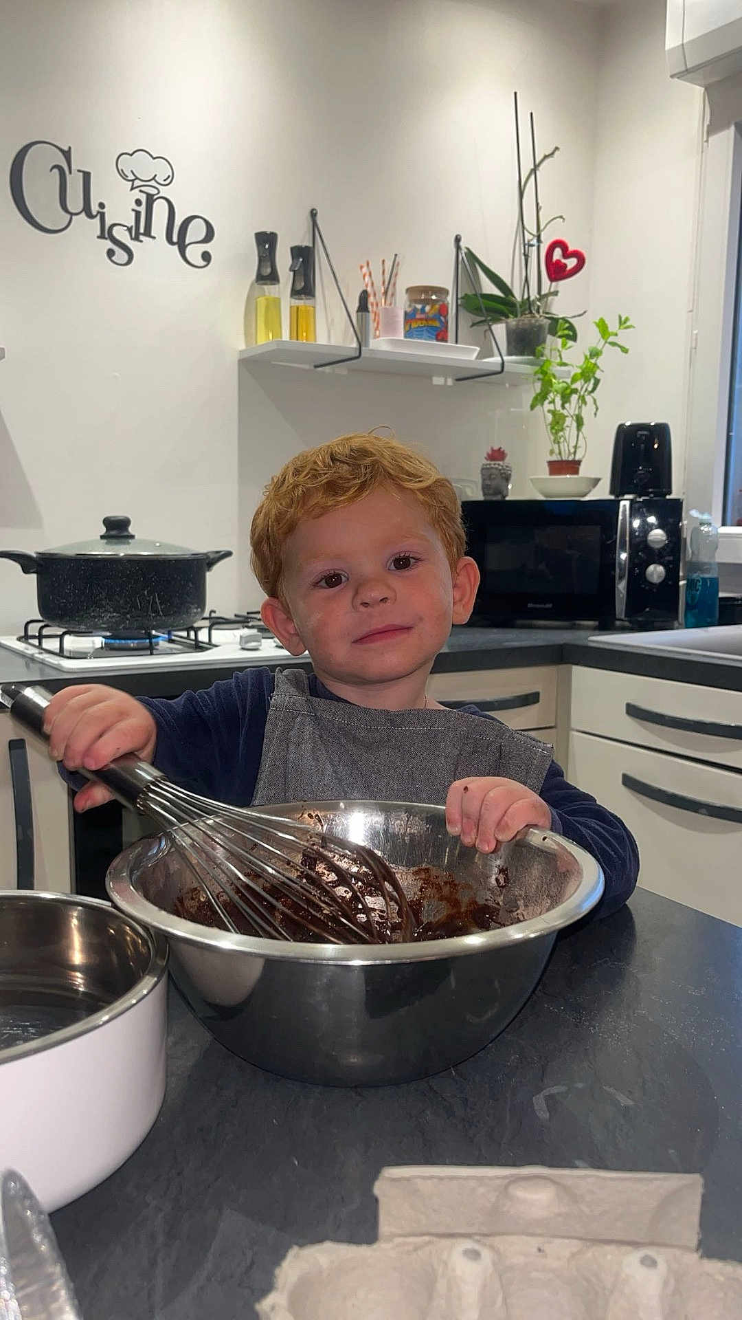 Mayrone participe au concours pour gagner de l'argent avec cette photo : child, toddler, kitchen, mixing_bowl, whisk, cooking, chocolate, apron, countertop, oven, pots, plants, microwave, cupboard, smile, red_hair, indoor, food_preparation, face, person