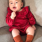Nyla is registered to the contest to win money with this photo: baby, infant, smiling, red_dress, headband, orange_socks, couch, soft_texture, happy, child, cute, indoors, portrait, sitting, skin, face, hand, legs, cozy, young