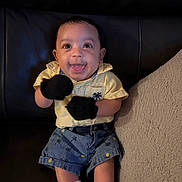 Ozias is registered to the contest to win money with this photo: baby, infant, smiling, sitting, leather_couch, pillow, socks, mittens, yellow_shirt, denim_shorts, pineapple_print, palm_tree_pattern, happy, tongue_out, big_eyes, portrait, indoor, adorable, small_child, hands