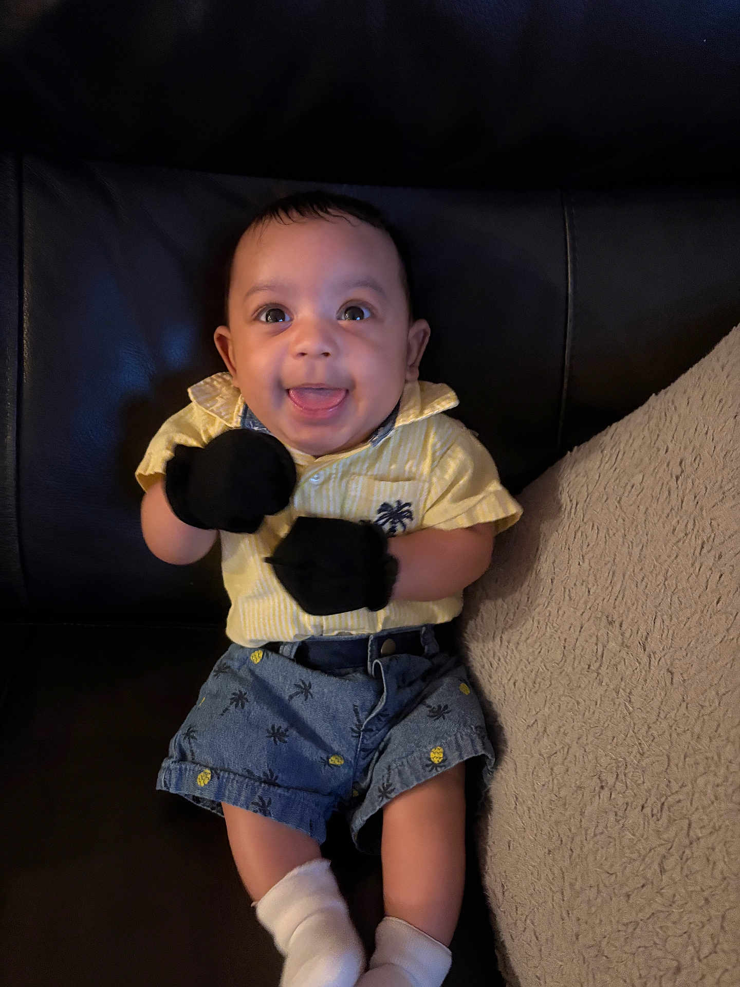 Ozias is registered to the contest to win money with this photo: baby, infant, smiling, sitting, leather_couch, pillow, socks, mittens, yellow_shirt, denim_shorts, pineapple_print, palm_tree_pattern, happy, tongue_out, big_eyes, portrait, indoor, adorable, small_child, hands