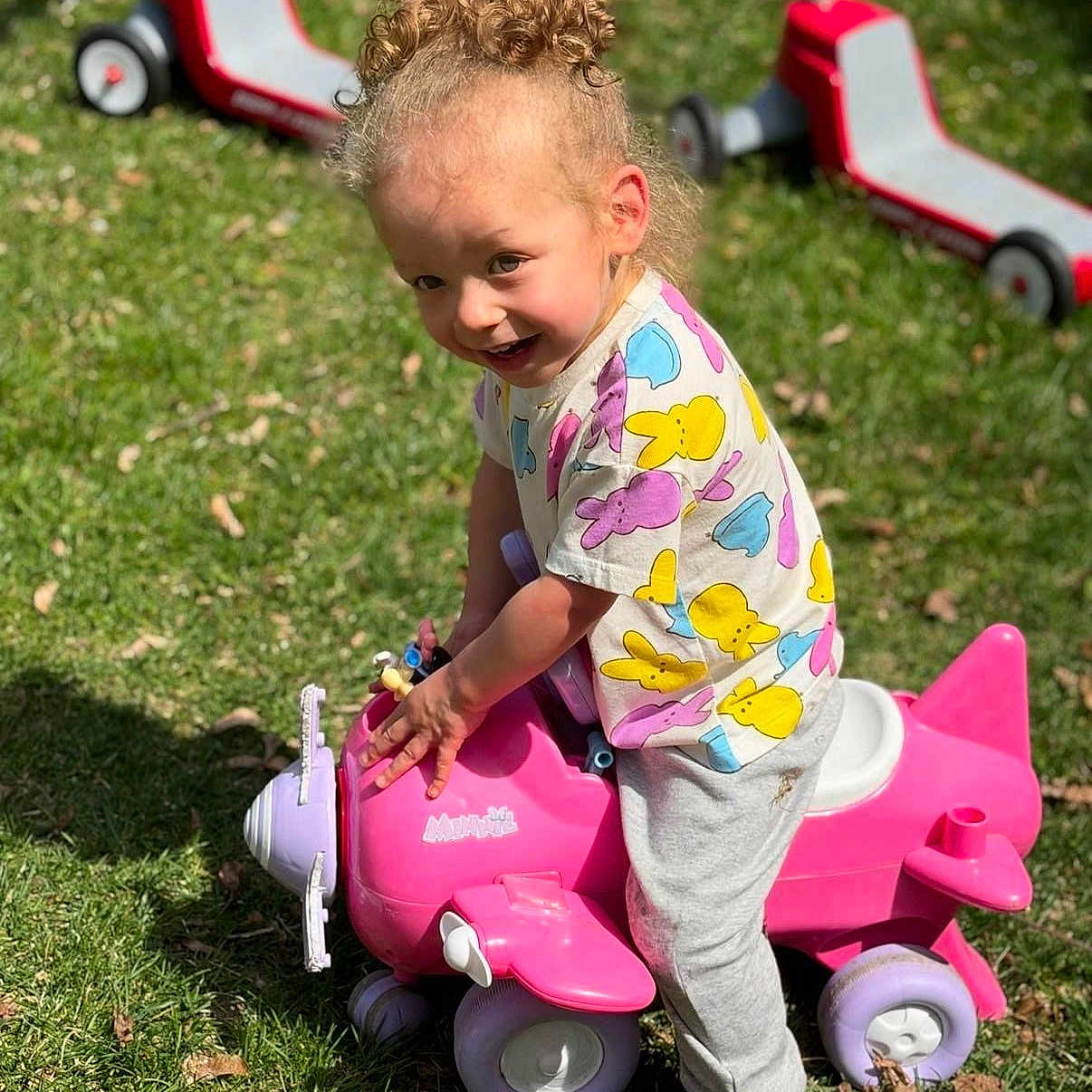 Amari is registered to the contest to win money with this photo: toddler, child, curly_hair, outdoor, grass, toy_airplane, pink, purple, scooter, play, sunny, shirt, bunny_pattern, pants, shoes, happy, smiling, daylight, person, fun