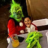 child, smiling, costume, holiday, creature, green, red, portrait, indoor, party, table, juice, jar, straw, phone, chair, clothing, festive, funny, photography