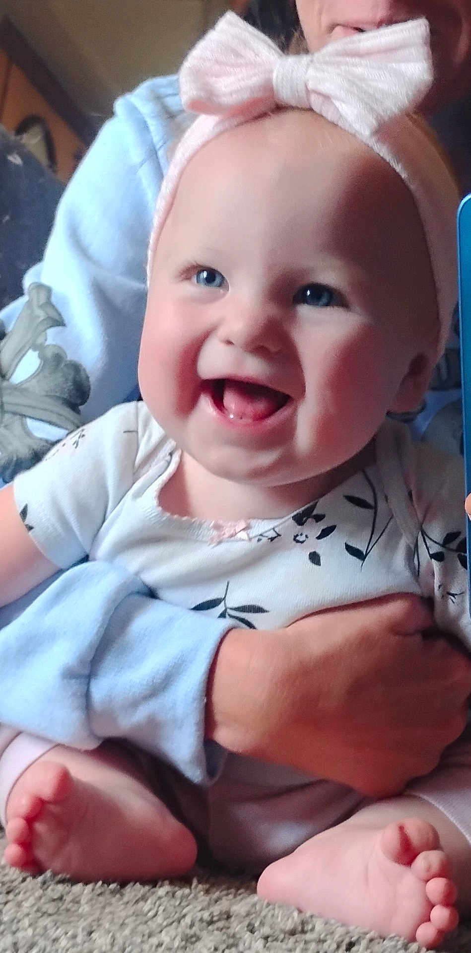 Raelynn is registered to the contest to win money with this photo: baby, smiling, blue_eyes, pink_bow, headband, infant, barefoot, carpet, toddler, adult_hand, hugging, cute, indoor, child, clothing, person, happy, closeup, sitting, portrait