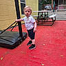basketball_hoop, bib, child, curious, daytime, fence, outdoor, person, picnic_table, playground, pole, red_floor, shirt, shorts, sneakers, standing, summer, toddler, trees, yellow_wall