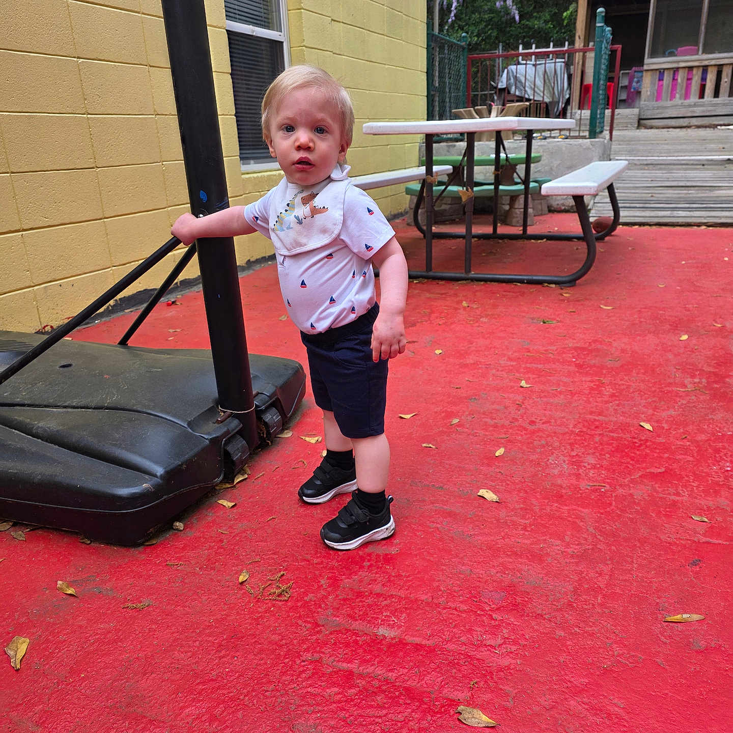 Nicolas joined the competition — help win amazing prizes! toddler, child, outdoor, basketball_hoop, pole, red_floor, yellow_wall, picnic_table, sneakers, bib, shorts, shirt, curious, standing, fence, trees, playground, summer, daytime, person