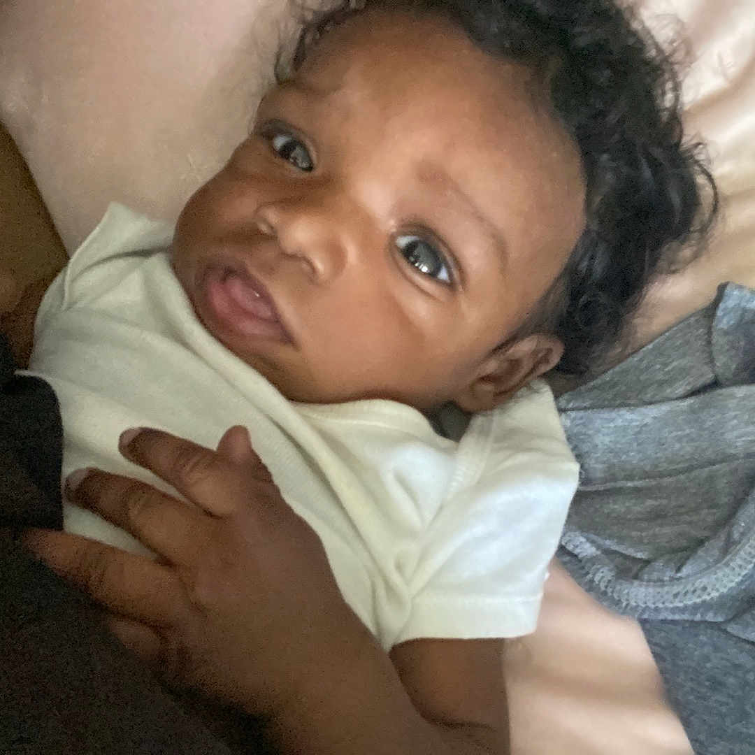 Noah is registered to the contest to win money with this photo: baby, bed, blanket, clothing, curly_hair, cute, fabric, face, hand, indoors, infant, lying_down, newborn, person, portrait, skin, sleepwear, soft_texture, white_shirt, young_child