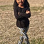 child, girl, smiling, outdoor, grass, sunlight, black_shirt, checkered_pants, sunflower_pattern, crocs, mismatched_socks, curly_hair, hair_bun, beads, standing, portrait, happy, playful_pose, casual_clothing, nature