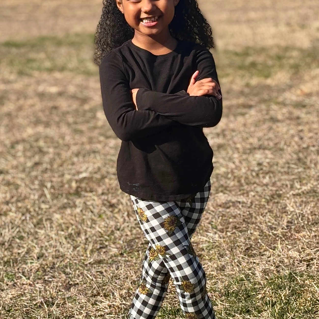 Peyton Carter joined the competition — help win amazing prizes! beads, black_shirt, casual_clothing, checkered_pants, child, crocs, curly_hair, girl, grass, hair_bun, happy, mismatched_socks, nature, outdoor, playful_pose, portrait, smiling, standing, sunflower_pattern, sunlight