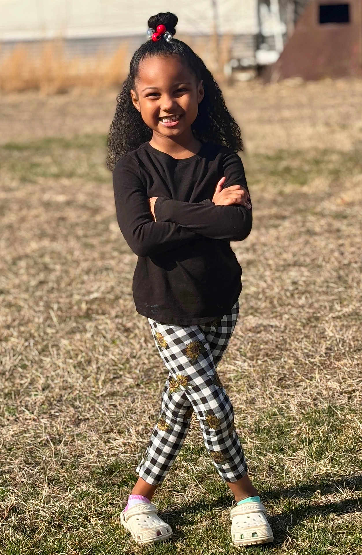 Peyton Carter joined the competition — help win amazing prizes! child, girl, smiling, outdoor, grass, sunlight, black_shirt, checkered_pants, sunflower_pattern, crocs, mismatched_socks, curly_hair, hair_bun, beads, standing, portrait, happy, playful_pose, casual_clothing, nature