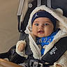 baby, child, smiling, stroller, jacket, headband, blue_clothing, jeans, footwear, indoor, seatbelt, happy, cute, portrait, person, infant, babywear, warm_clothing, face, hands