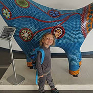 Roman is registered to the contest to win money with this photo: child, smiling, backpack, curly_hair, mosaic, sculpture, colorful, indoor, art, pattern, standing, happy, casual_clothing, shoes, floor, sign, exhibit, playful, person, museum