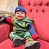 toddler, child, smiling, red_chair, hoodie, cap, boots, indoor, cozy, plush_furniture, happy, casual_clothing, portrait, seated, cute, young_child, fashion, colorful, relaxed, face