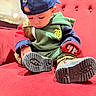 toddler, child, cap, hoodie, shoe, sitting, red_couch, indoor, curious, footwear, casual_clothing, person, young_child, close_up, seated, colorful_clothing, looking_down, soft_furniture, candid, portrait